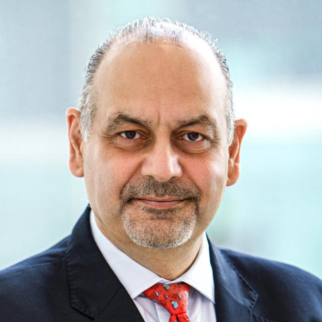 A man in a formal suit and patterned tie stands in front of a blurred urban background, exuding professionalism and confidence.