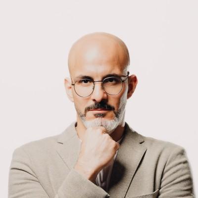 Bald man wearing a beige suit jacket with his hand resting thoughtfully on his chin against a plain light background.